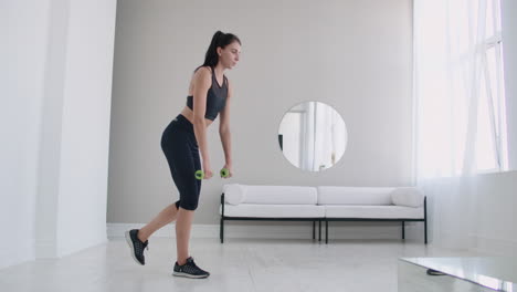 Young-beautiful-brunette-woman-standing-on-one-leg-doing-bends-with-dumbbells.-Performing-deadlift-with-dumbbells-at-home.